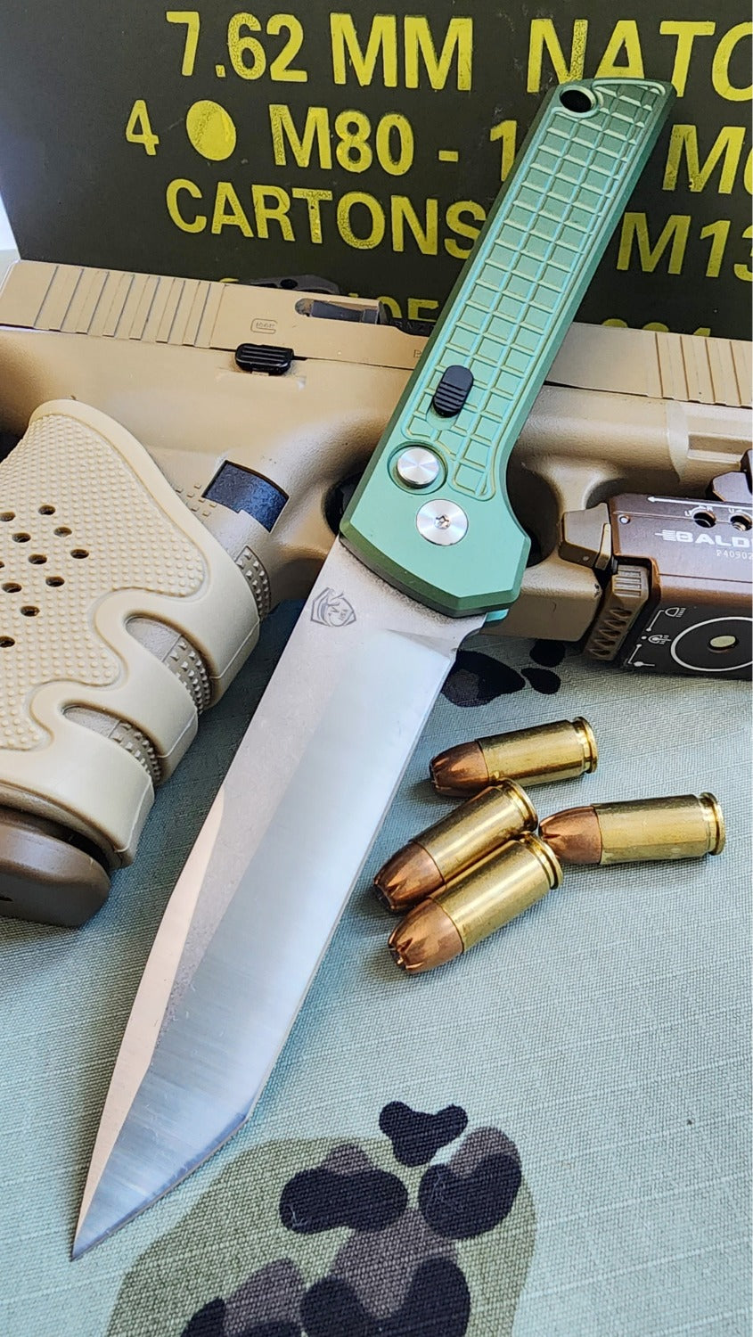 Knife with green handle and silver blade on a surface with ammunition and tactical equipment in the background.
