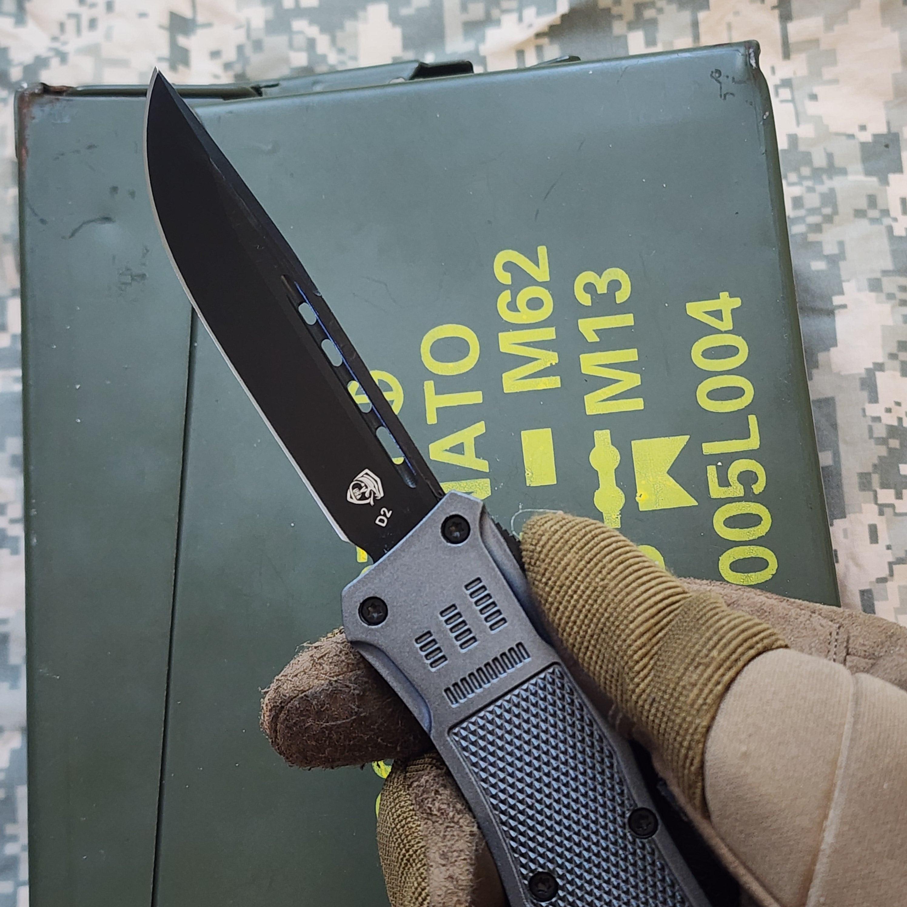 Folding knife held by a gloved hand with a military-style box in the background