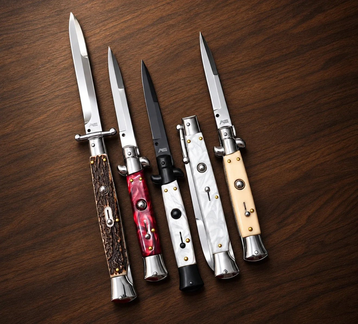 Five stiletto and swinguard switchblade knives with stag-style, red pearl, white pearl and ivory-colored handles on wood.