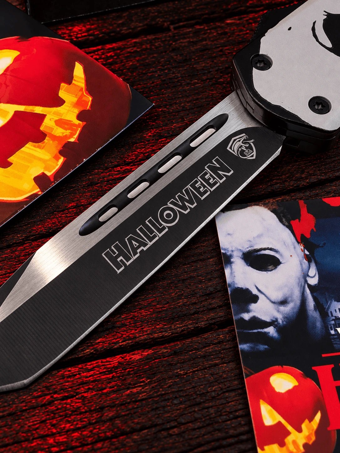 A Halloween-themed knife with a metallic blade engraved with 'HALLOWEEN' and a skull handle, displayed on a dark wooden surface with Halloween-themed cards.