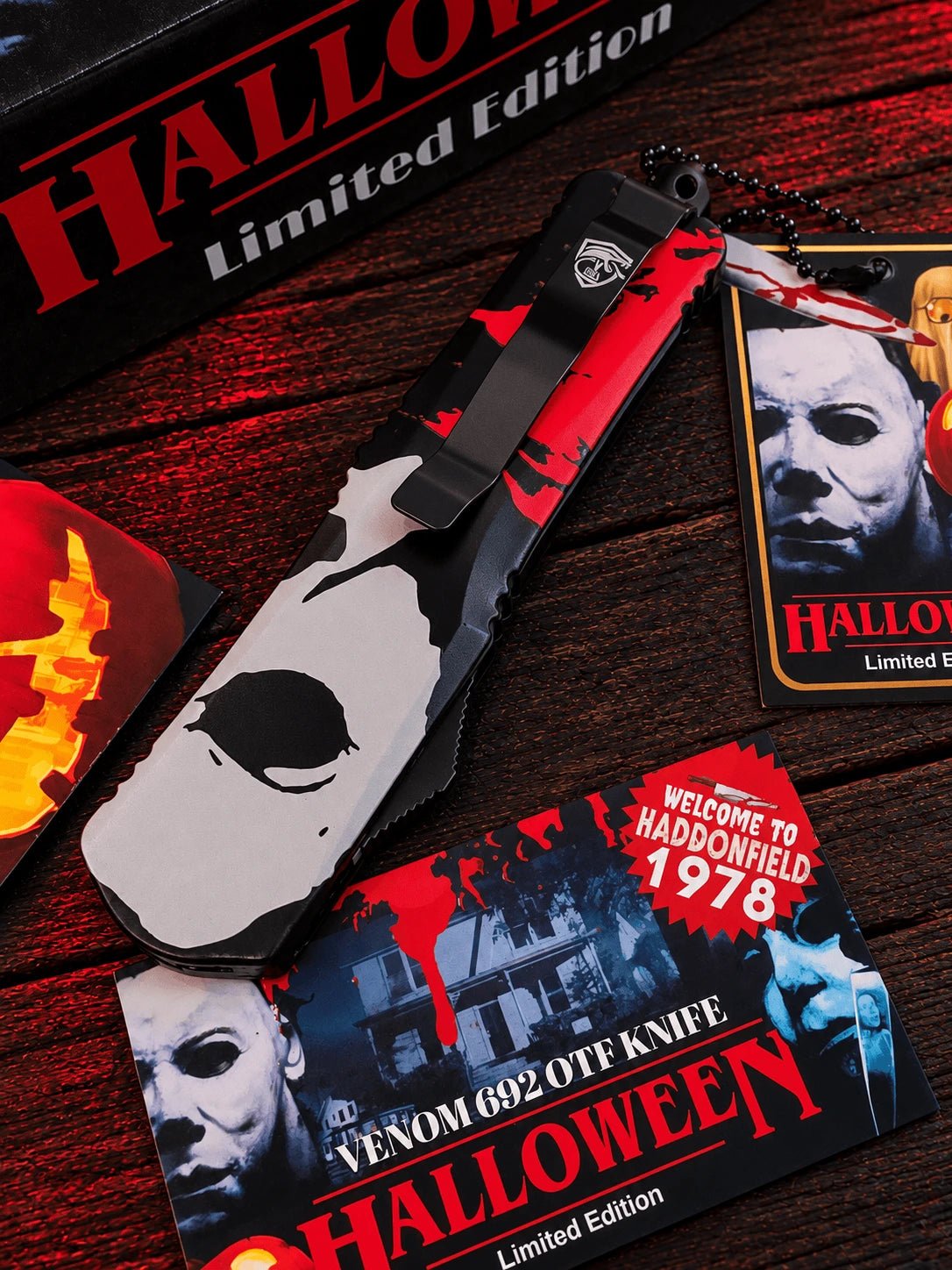 A Venom 092 OTF Knife from the Halloween Limited Edition, featuring a black and white skull design with red blood splatters, displayed on a dark wooden background alongside themed packaging and accessories.